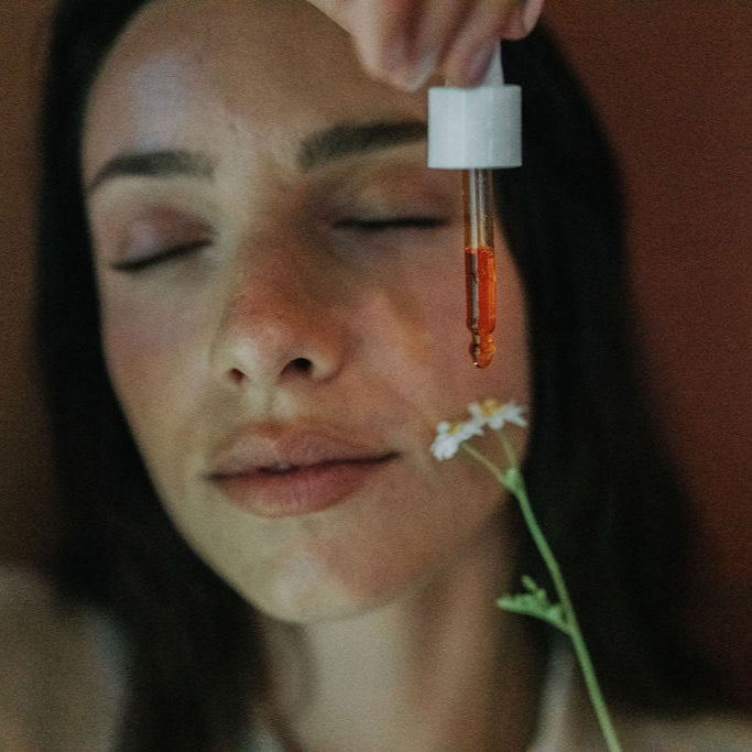 Woman with eyes closed, dropper with orange liquid held above her face, blurred background