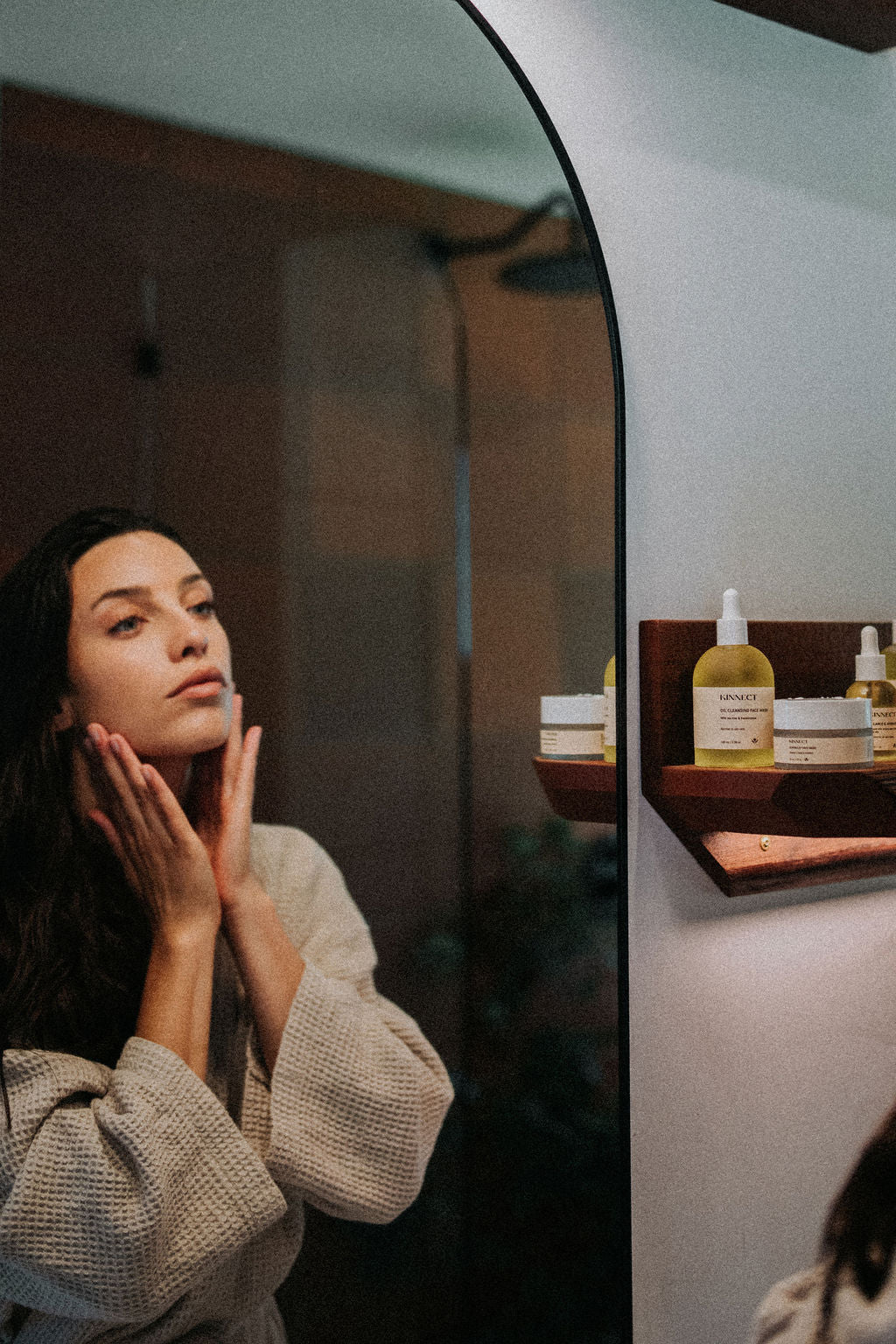 Woman applying skincare product in front of a mirror with skincare products on a shelf.