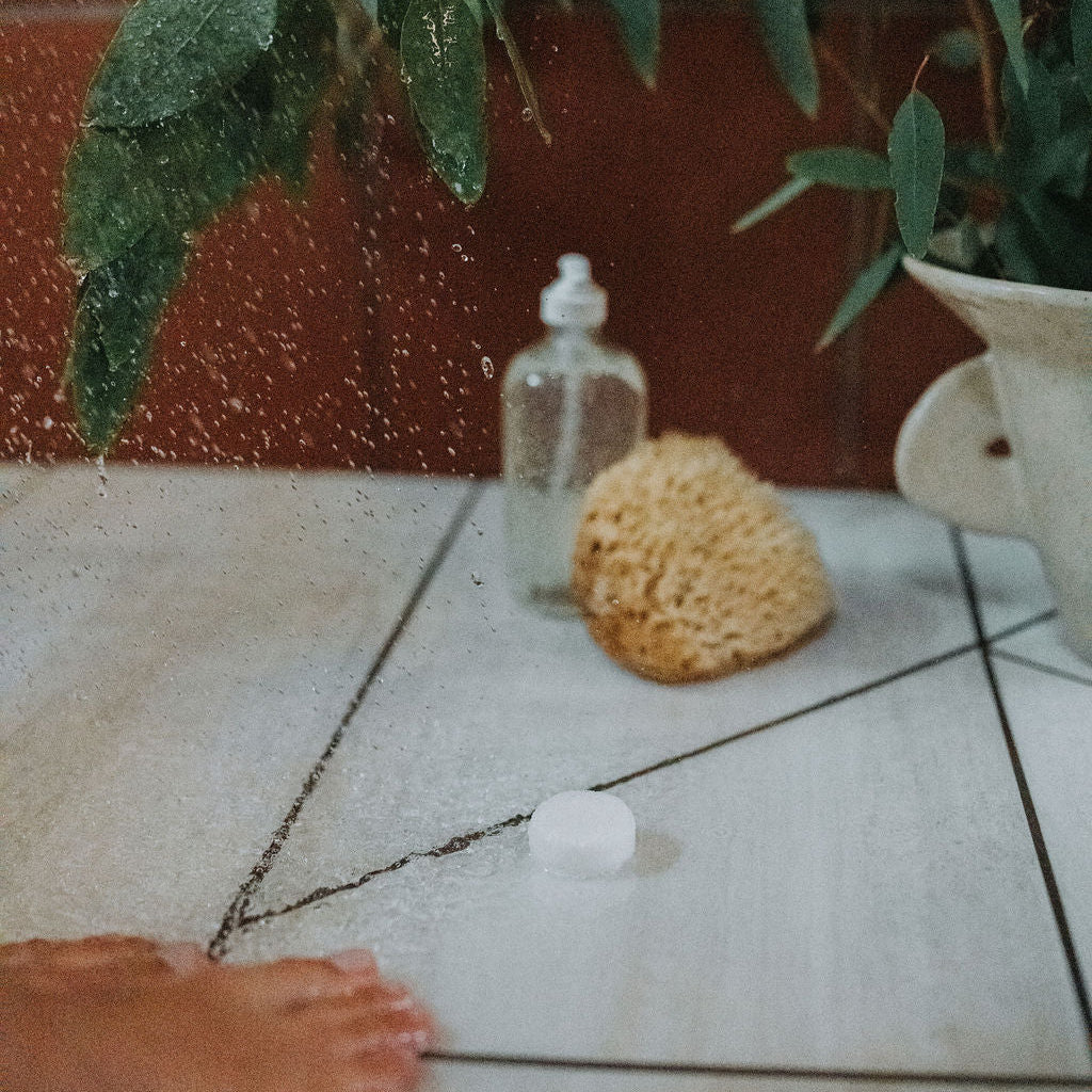 Foot on a tiled floor with a bottle, sponge, and plant in the background. A shower steamer sits in the centre.