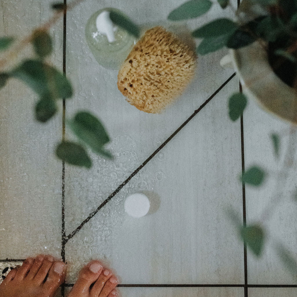 Person's feet on a tiled floor with a glass of water, sponge, and white shower steamer.