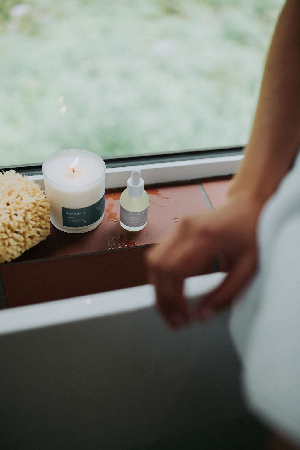 Kinnect Candle, bottle, and sea sponge on a windowsill with a blurred person in the foreground.