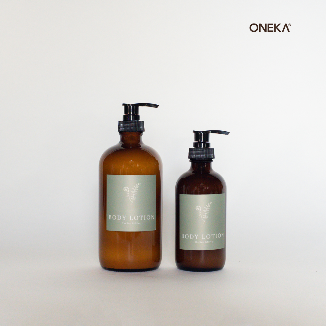 Two amber glass bottles filled with ONEKA cedar and sage body lotion sit side by side on a glass surface. One is 500ml (larger, left), and the second is 250ml (smaller, right). They both have a green Den Refillery label with an illustrated fern and text that reads, “Body Lotion”