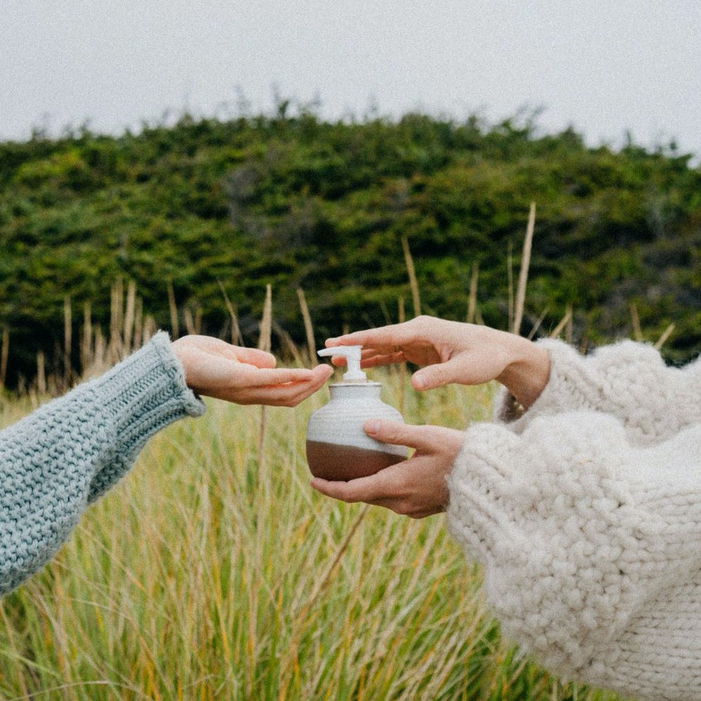 Two people, who are just out of frame and wearing beautiful poofy hand-knit sweaters, pump body lotion into each others hands with one of the Kay ceramics ceramic soap dispensers. There is beautiful long grass behind them with a coastal bush even further in the background