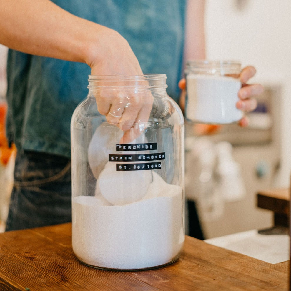 Clear 4 Litre Glass jar with hydrogen peroxide in it. A Den Refillery employee is Refillery their smaller glass jar with the bulk in the 4 Litre glass jar