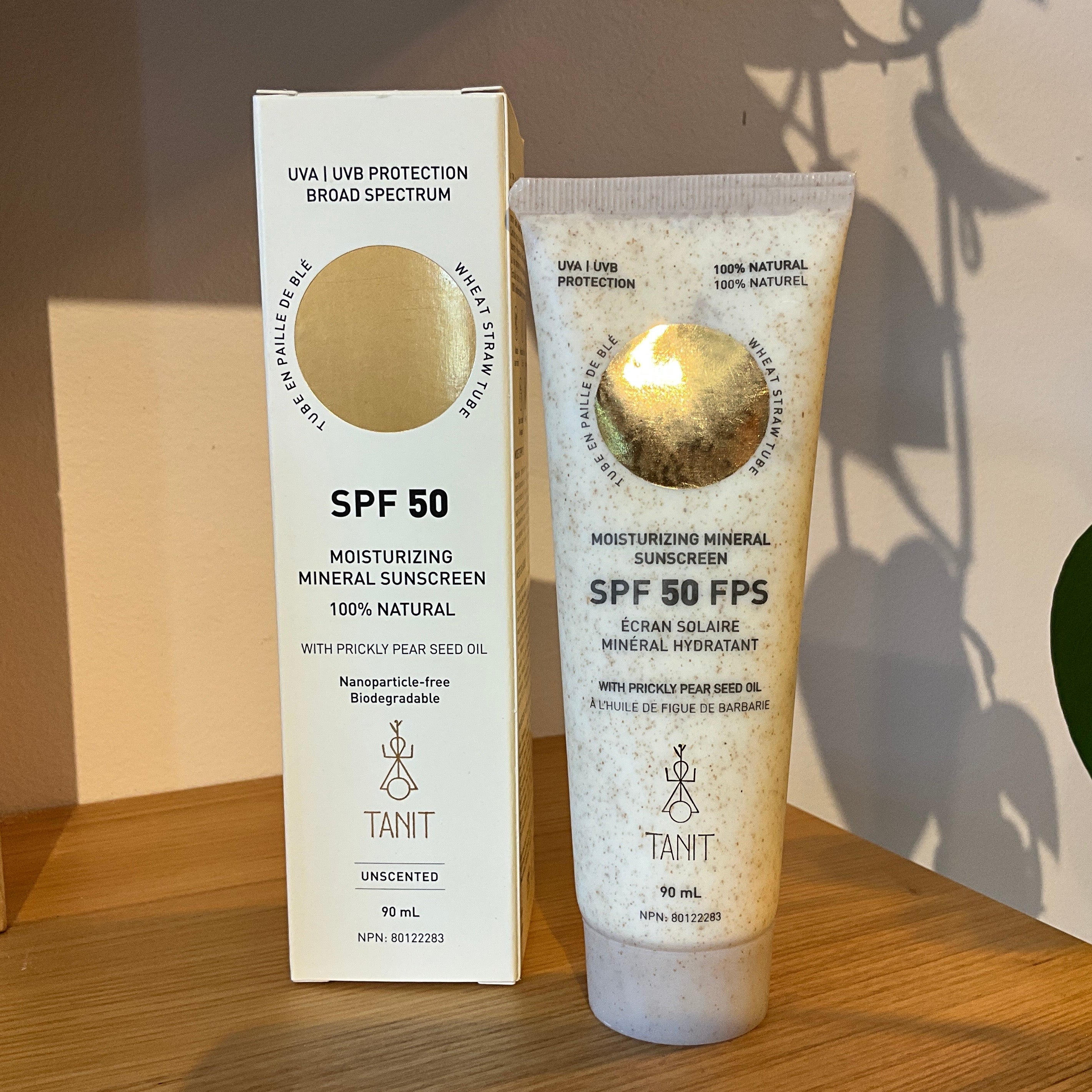 Taint SPF 50 sunscreen shown with its white outer box and a clear squeeze tube containing a milky beige product, placed on a wooden surface with a plant leaf and wall shadows in the background.