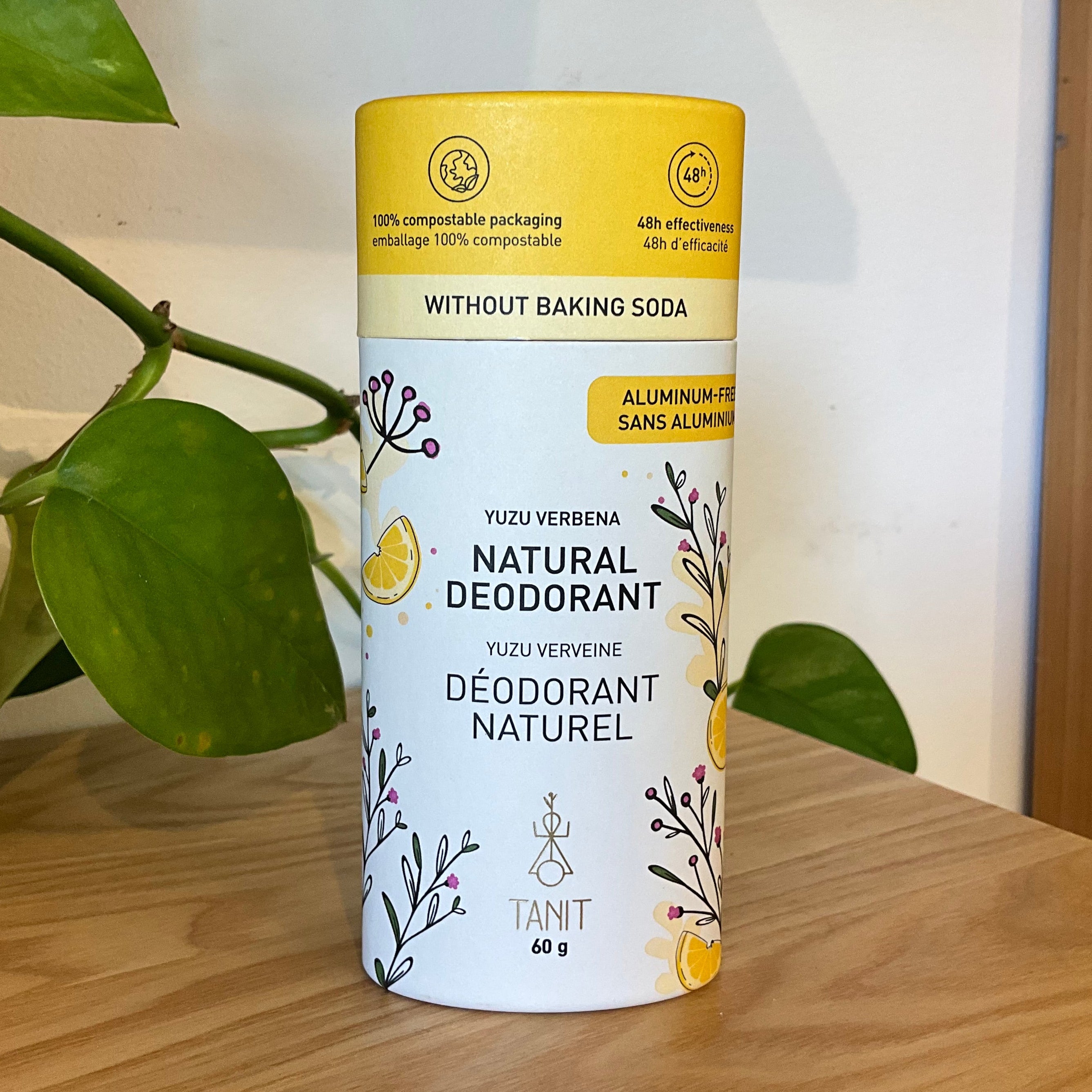 A cylindrical canister of Yuzu Keveren Natural Deodorant by TANT, placed on a wooden table with green houseplant leaves in the background.