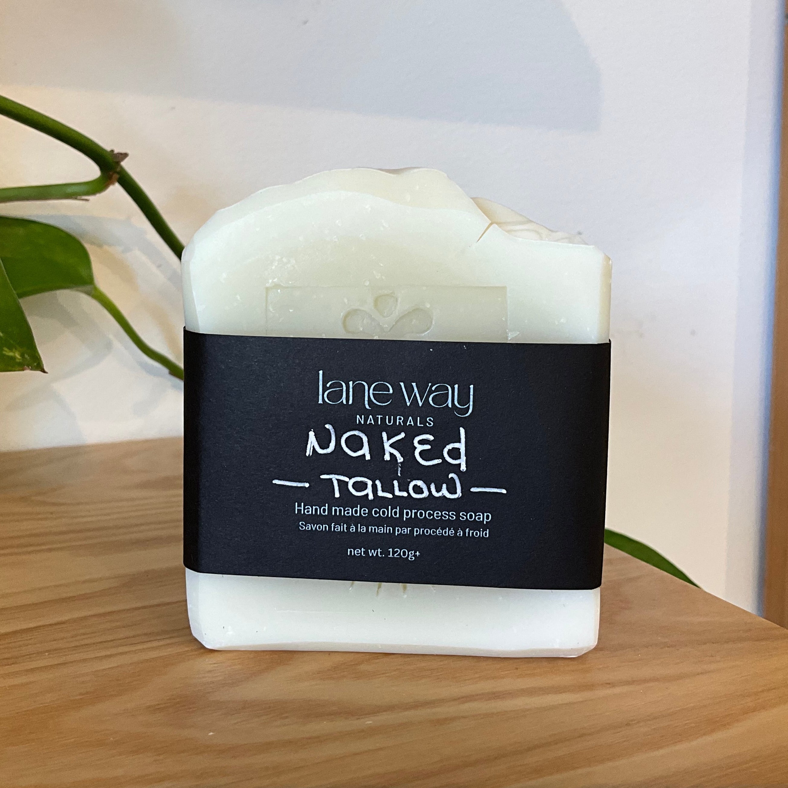 A bar of Lane Way Naturals Naked Tallow hand made cold process soap, wrapped in a black paper label, displayed on a wooden surface with a green plant leaf in the background.