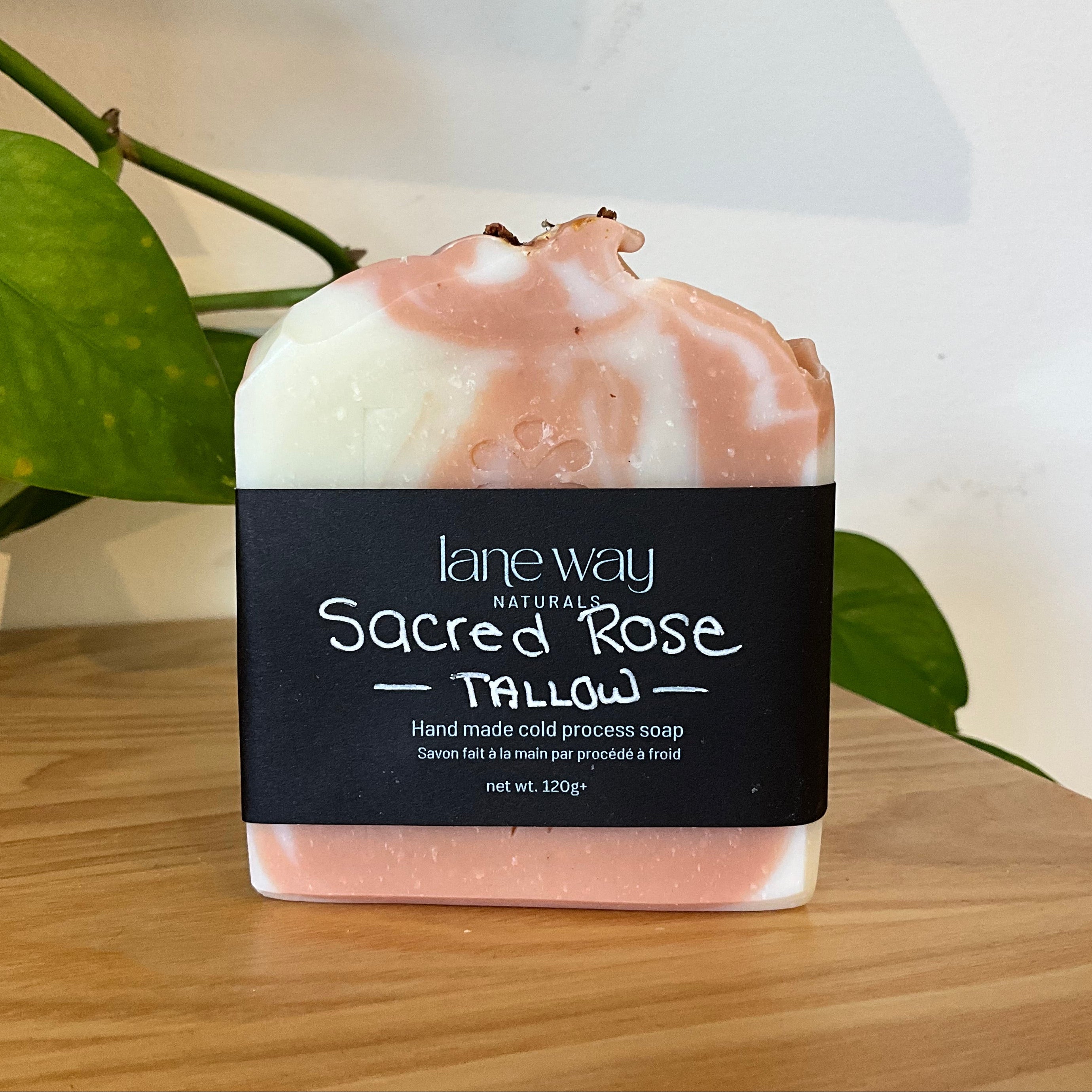 A handcrafted bar of soap with a black label reading 'lane way Natural Sacred Rose Tallow' sits on a wooden surface with a green plant leaf in the background.