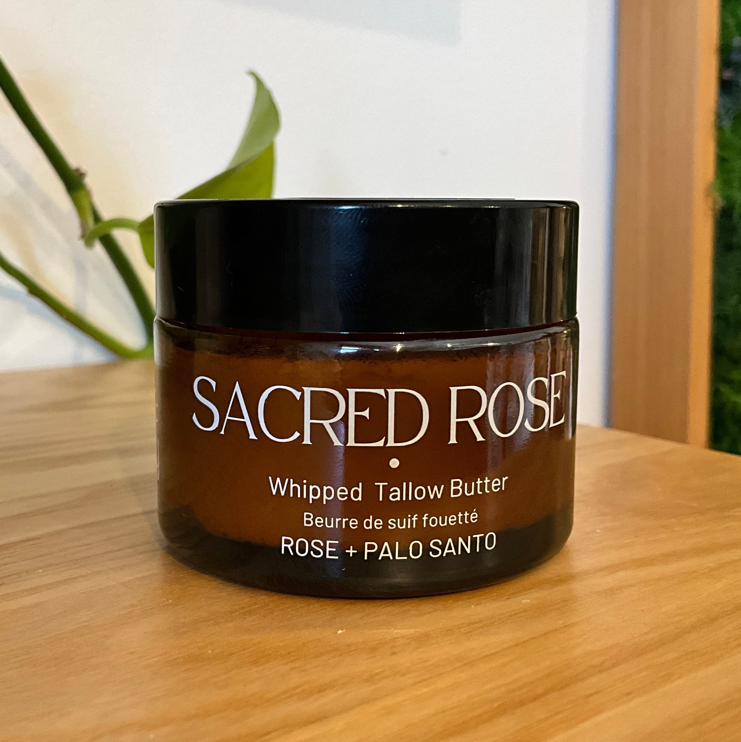 A jar of Sacred Rose Whipped Tallow Butter sits on a light wooden table. The product is in a dark amber brown jar with a black lid, featuring white text.