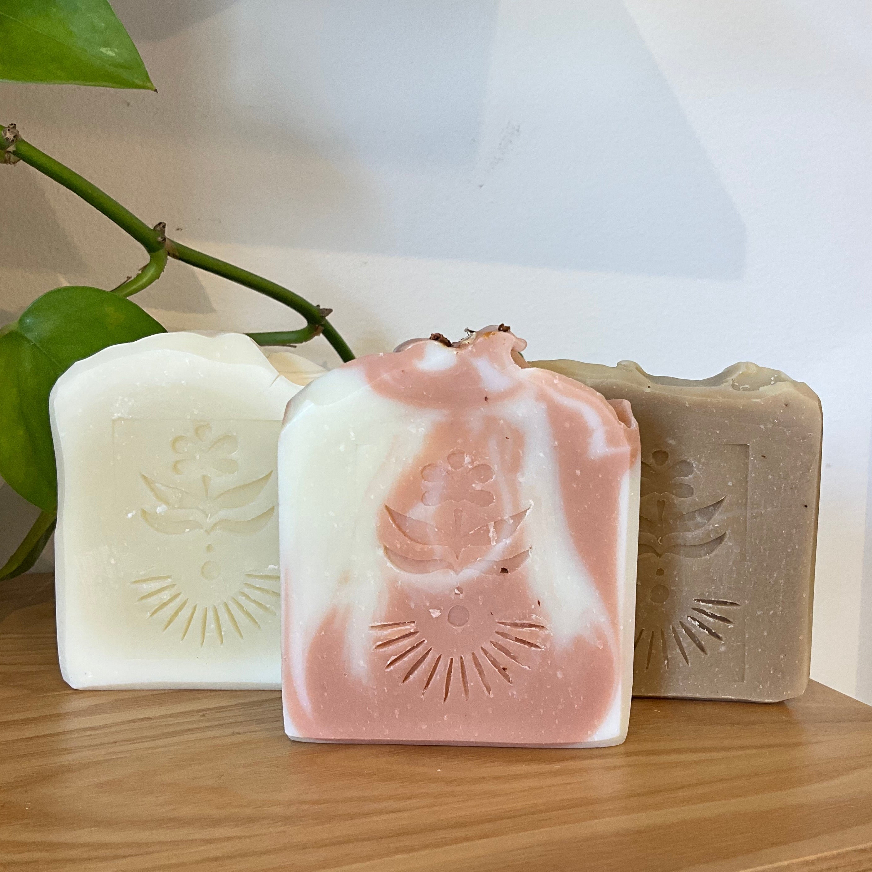 Three handmade rectangular bar soaps displayed on a wooden shelf, featuring a white soap with an embossed floral design, a pink and white swirled soap with a sunburst design, and a textured gray soap with an embossed eye design.