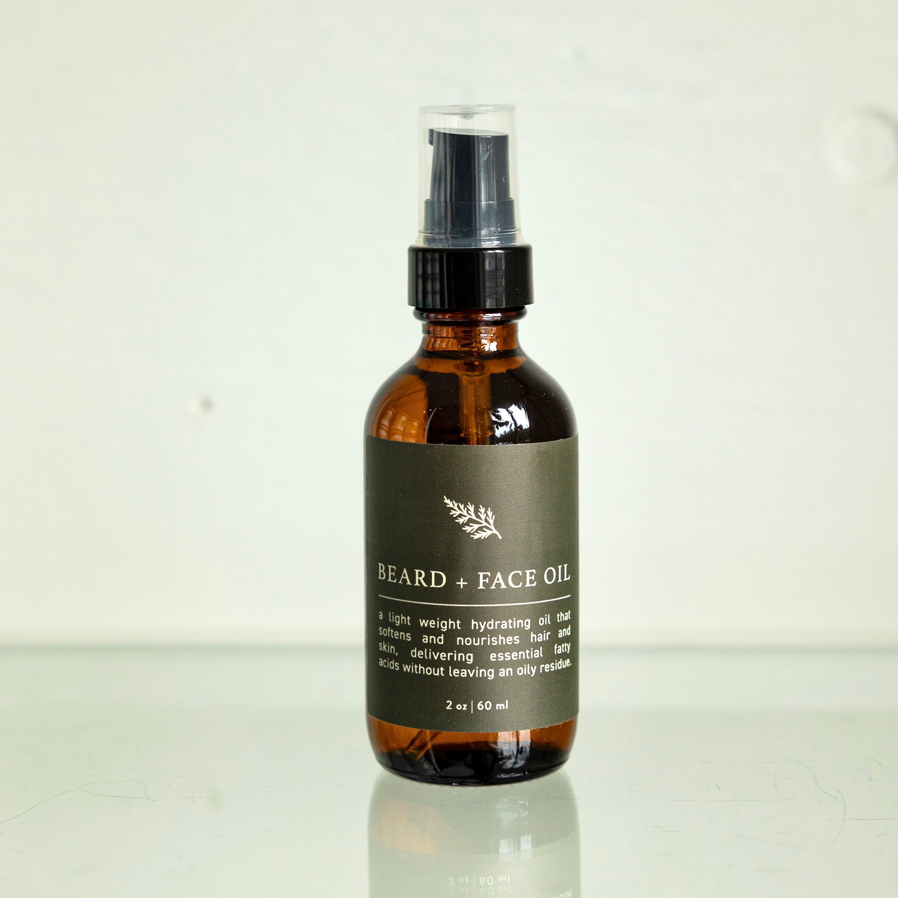 The Den Refillery Beard + Face Oil sits on a glass surface with a white background. The amber glass bottle features a black label with a cedar branch and reads, “A light weight hydrating oil that softens and nourishes hair and skin, delivering essential fatty acids without leaving an oily residue.”