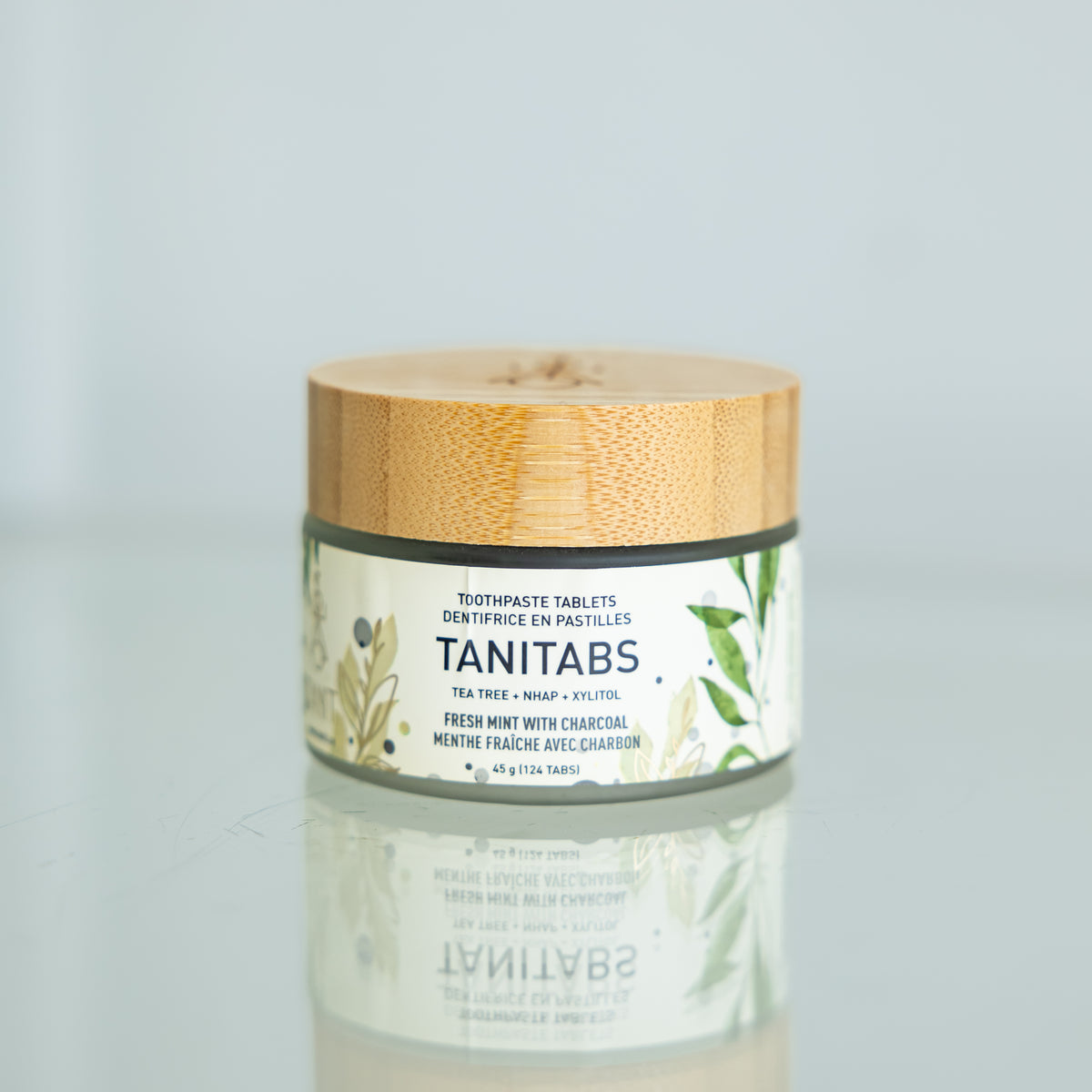 A glass jar with a bamboo lid containing toothpaste tablets, labeled TANITABS, with a reflection on a shiny surface.