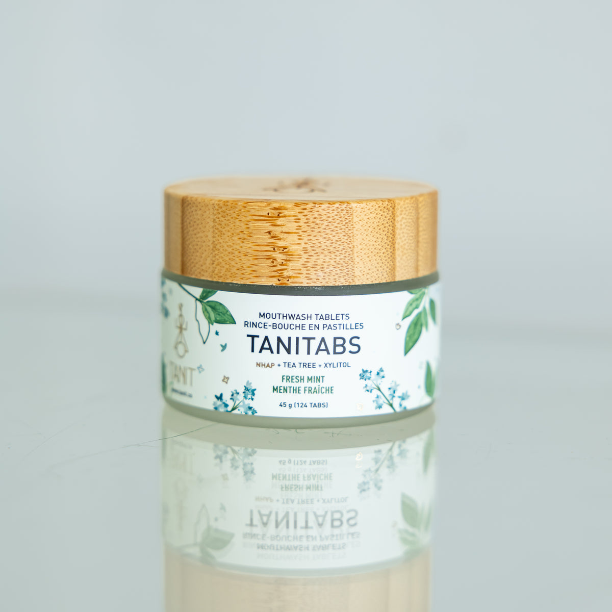 A glass jar with a bamboo lid containing toothpaste tablets, labeled TANITABS, with a reflection on a shiny surface.