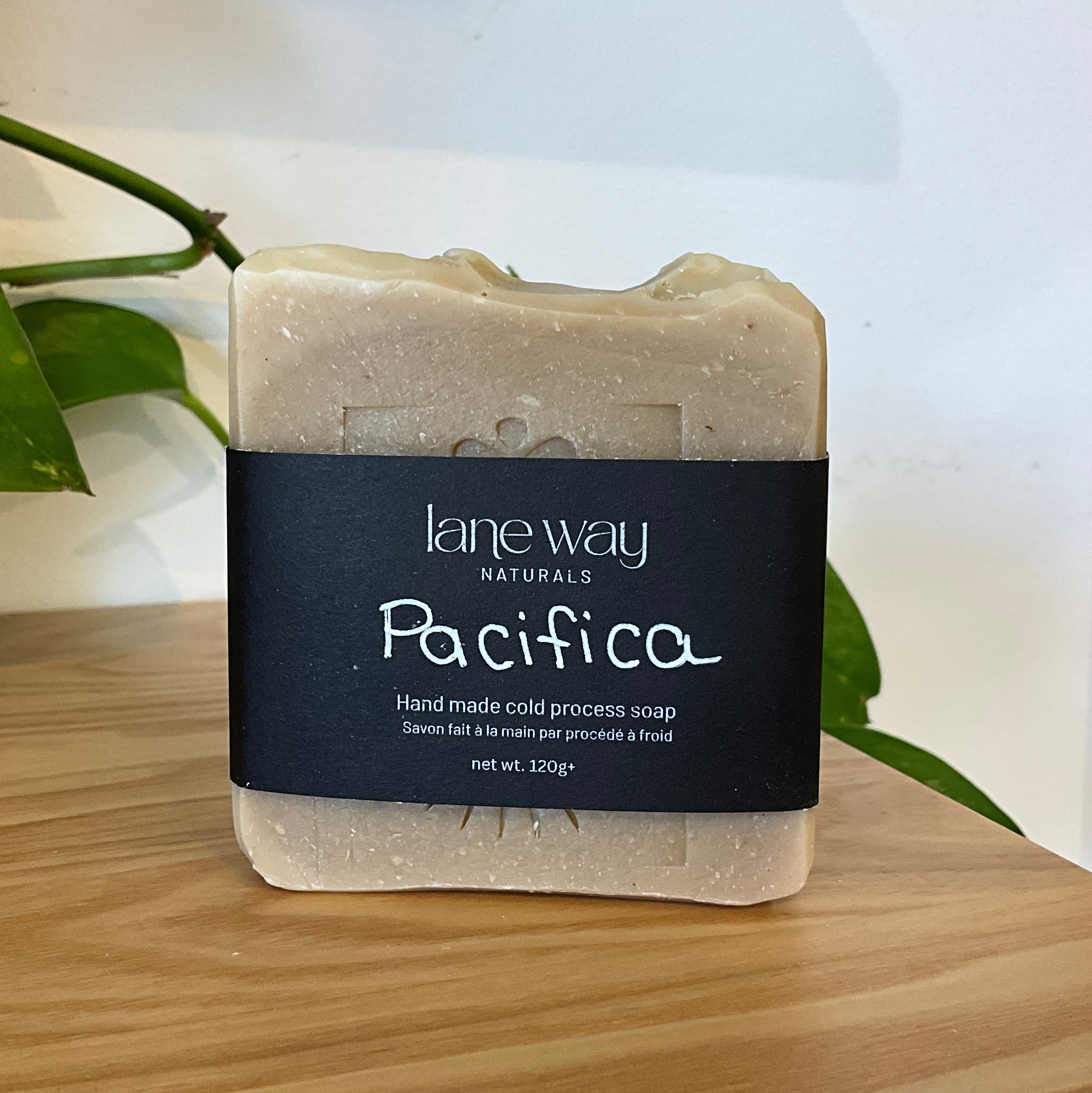 A bar of Lane Way Naturals 'Pacifica' handmade cold process soap, wrapped in a black paper label, displayed on a light wooden surface with a green plant leaf in the background.
