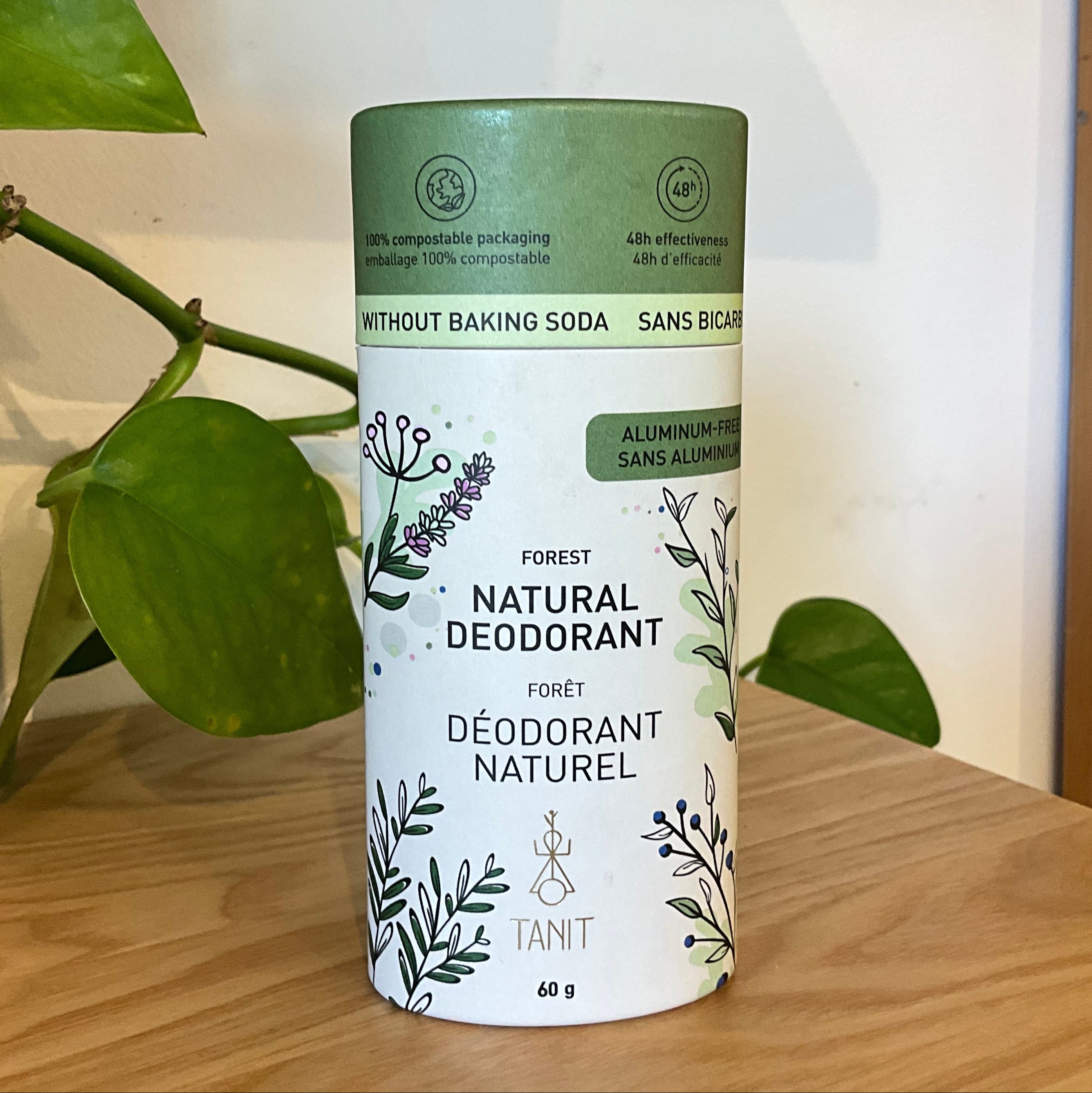 A 60g canister of TANIT Forest Natural Deodorant standing on a wooden shelf, flanked by a green houseplant and a wooden frame.
