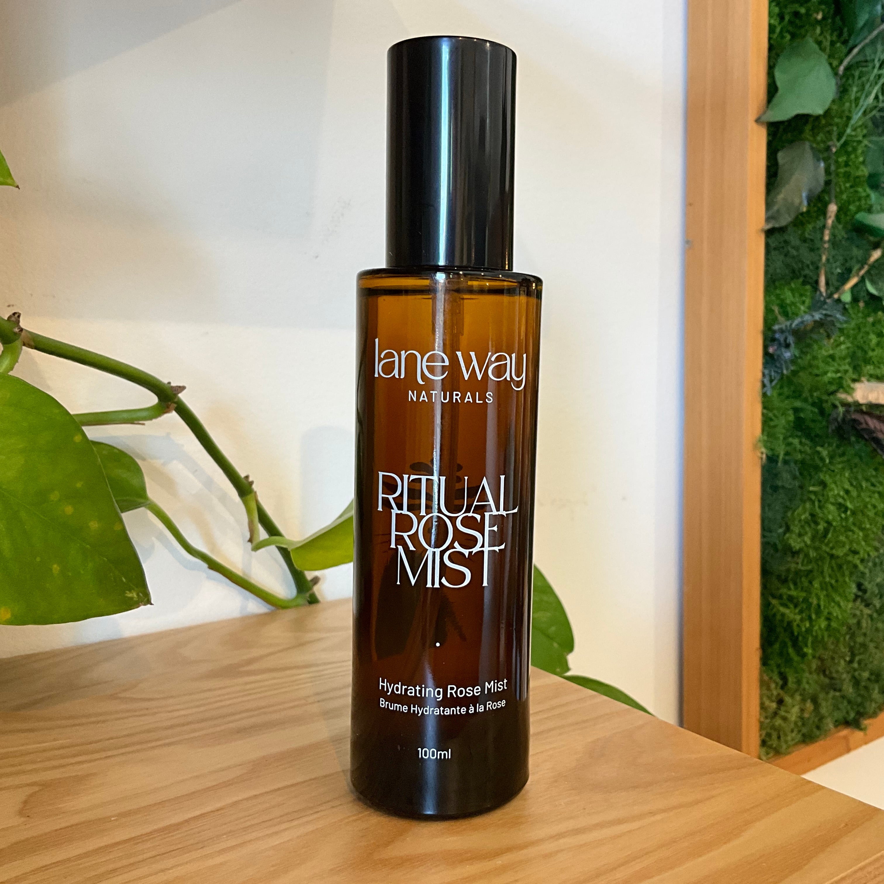 Lane Way Naturals Ritual Rose Mist in a dark amber glass bottle with a black cap, displayed on a wooden surface next to a green plant and a moss wall.
