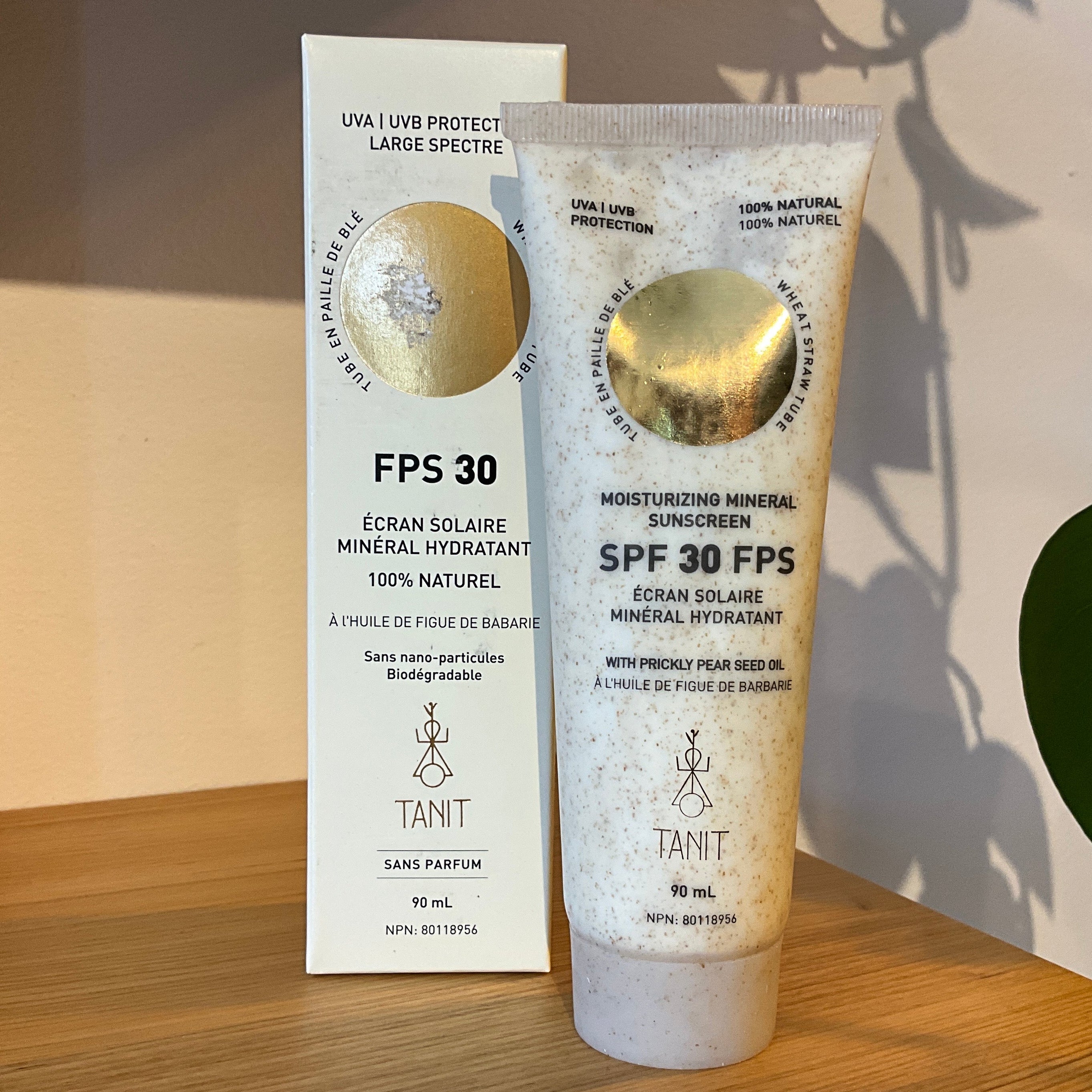 Tant FPS 30 Moisturizing Mineral Sunscreen shown as a white squeeze tube with a gold circular window next to its matching white cardboard box, displayed on a wooden shelf with a green plant leaf in the foreground.