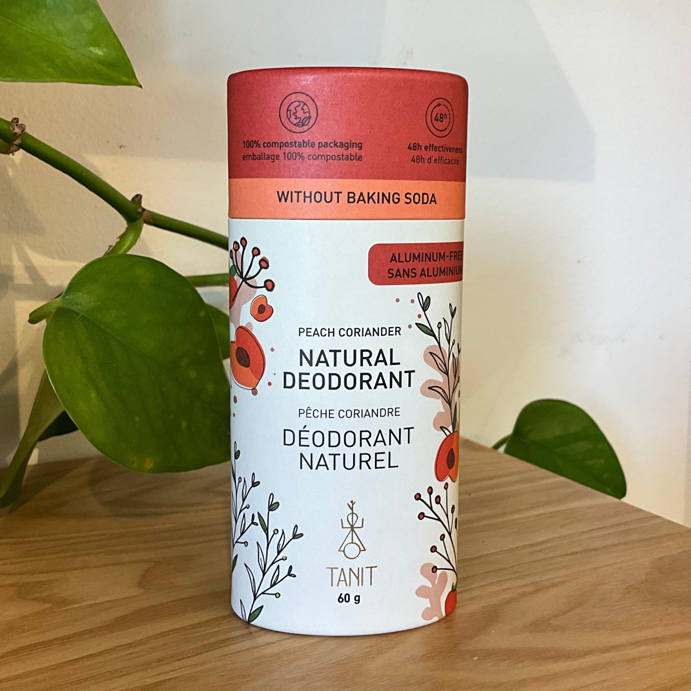 A cylindrical tin of TANT Natural Deodorant in Peach-Corssander scent, placed on a wooden table with a green plant in the background.