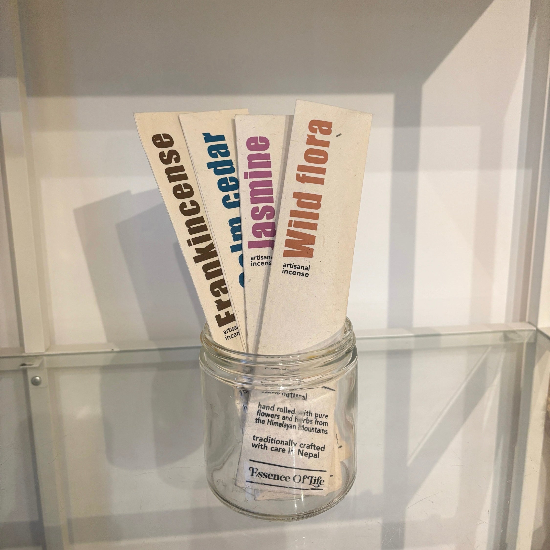 Jar with incense sticks labeled 'Frankincense', 'Cedar', 'Jasmine', and 'Wild Flora' on a reflective surface.