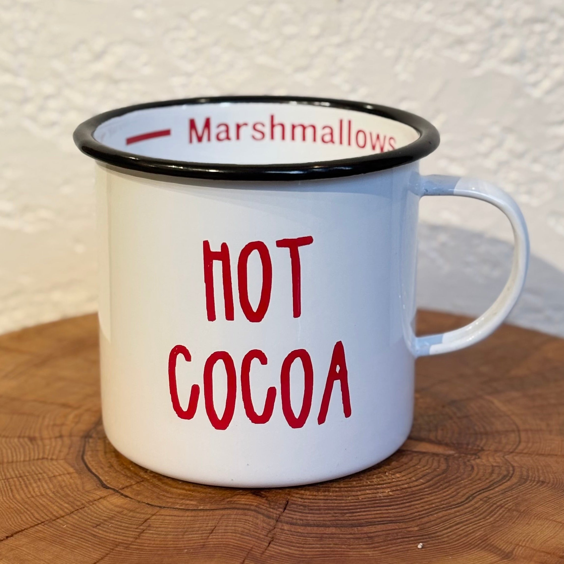 White mug with red text 'Hot Cocoa' on a wooden surface with a textured white wall background