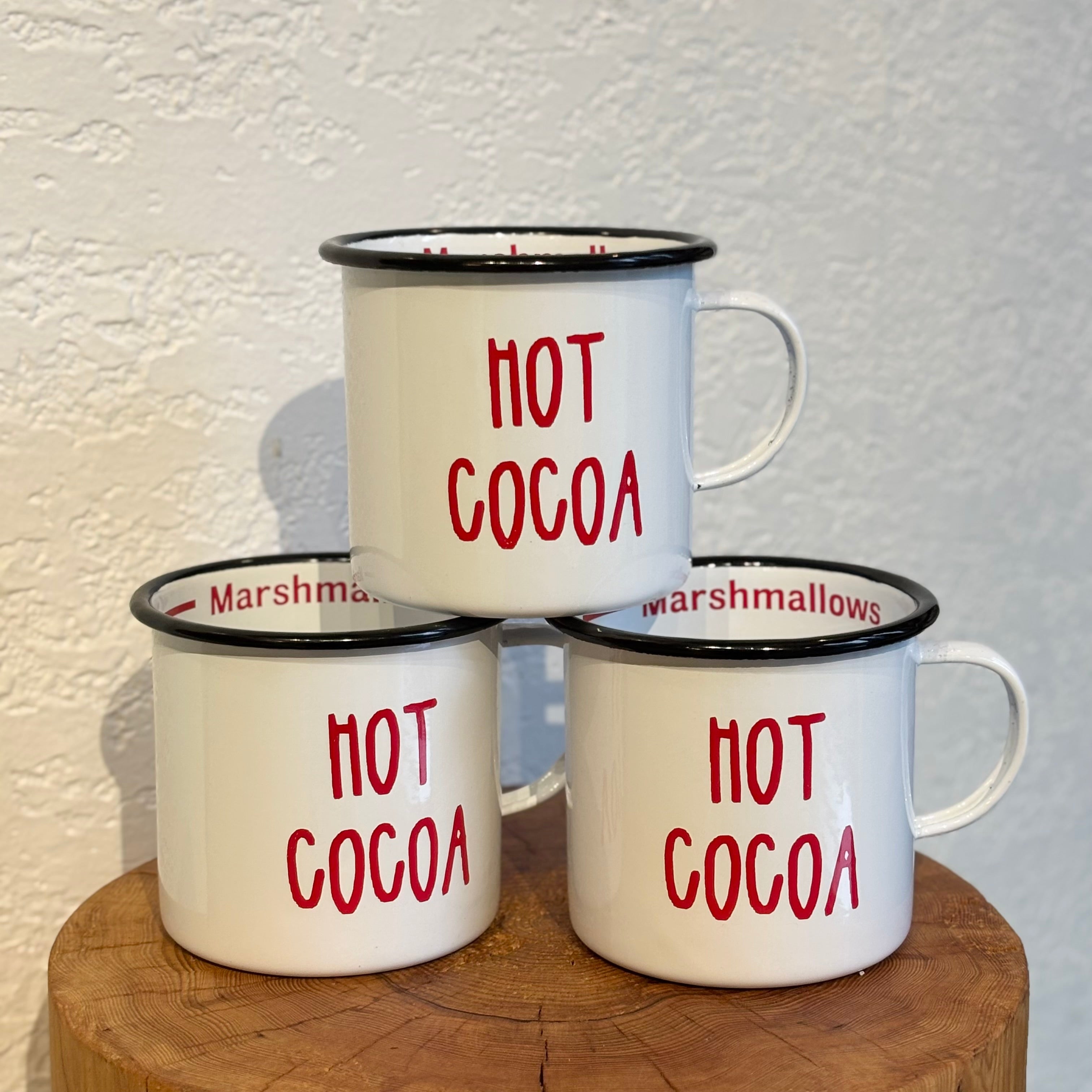 Three white mugs with red 'Hot Cocoa' text on a wooden surface against a light background