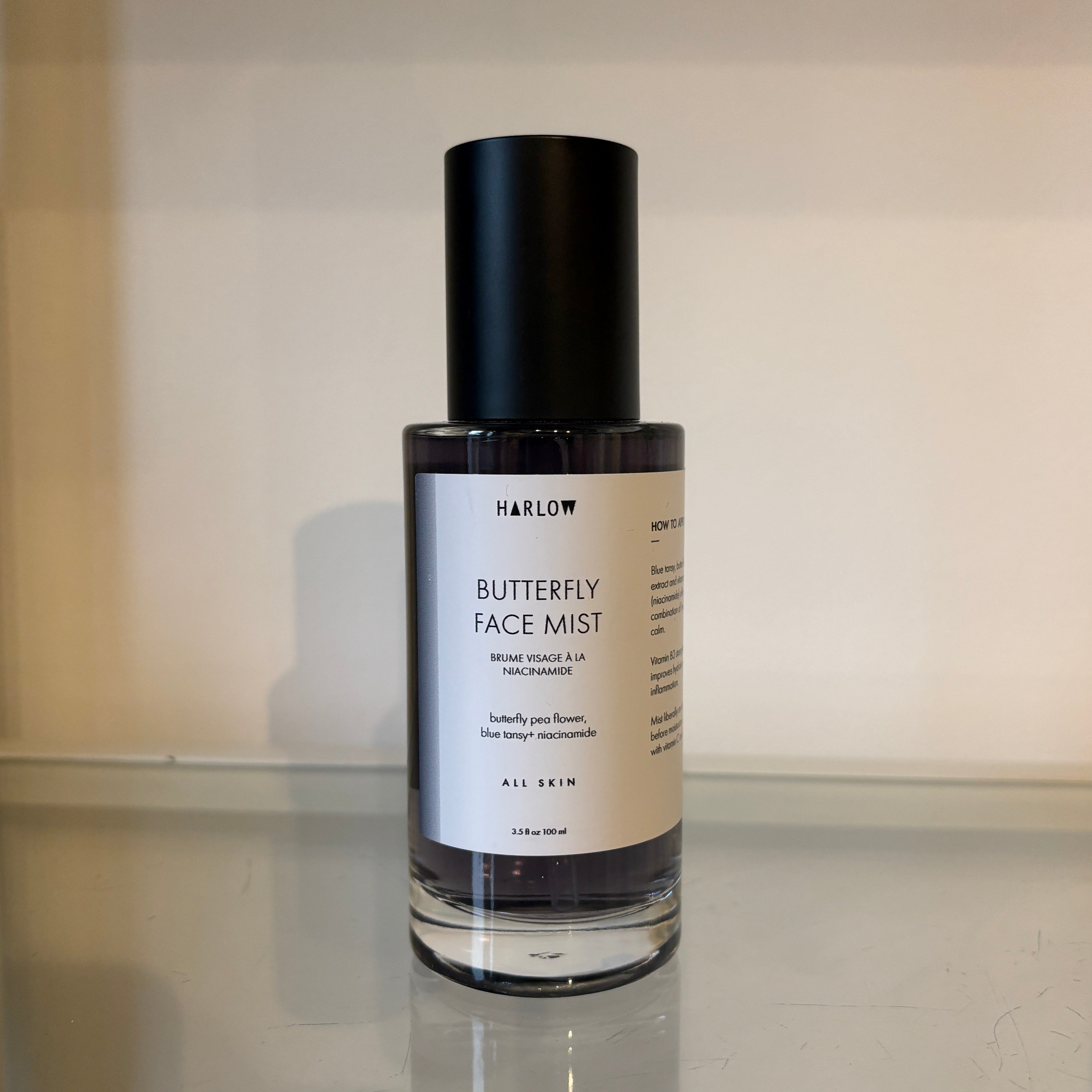 Butterfly face mist bottle with black cap and white label on a reflective surface.