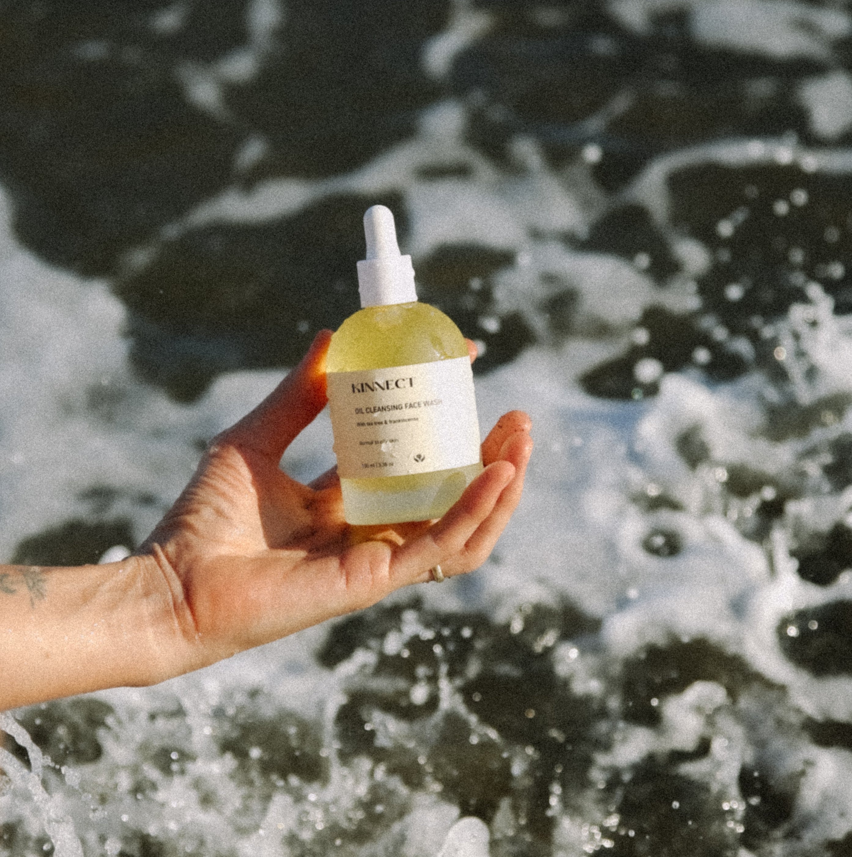Person holding a bottle of Kinnect Oil Cleanser product with ocean waves in the background