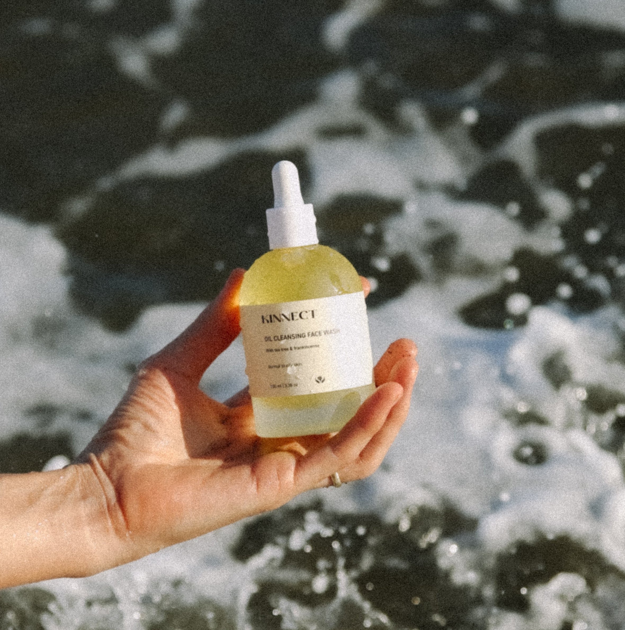 Person holding a bottle of Kinnect Oil Cleanser product with ocean waves in the background