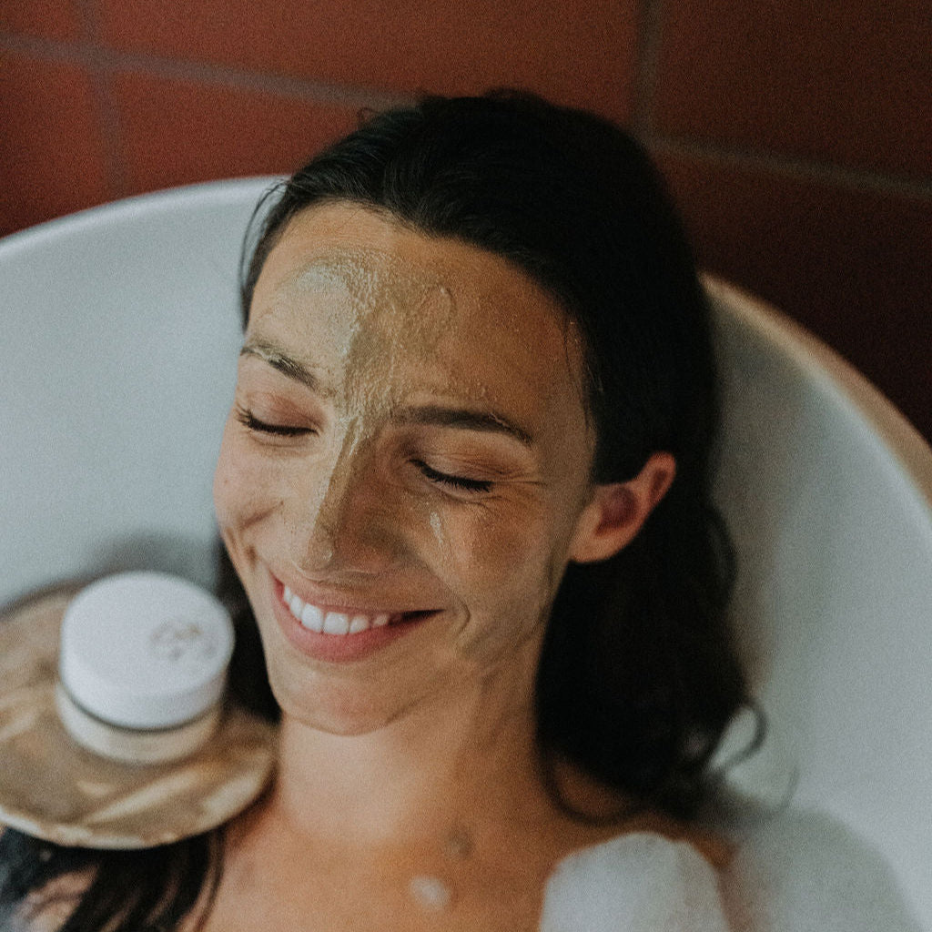 Woman in a bathtub with a facial mask on her face, surrounded by bubble bath bubbles.