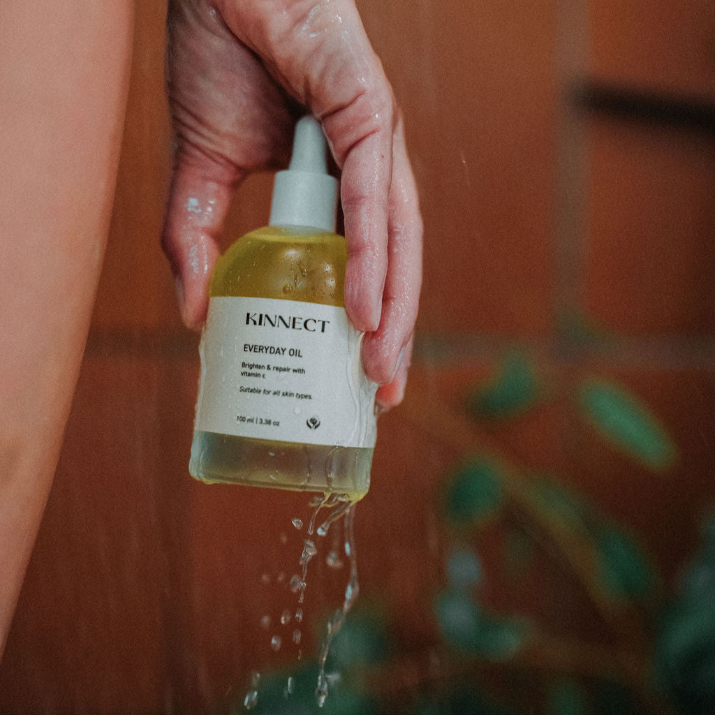 Hand holding a bottle of Kinnect skincare product with water droplets, against a tiled wall background.