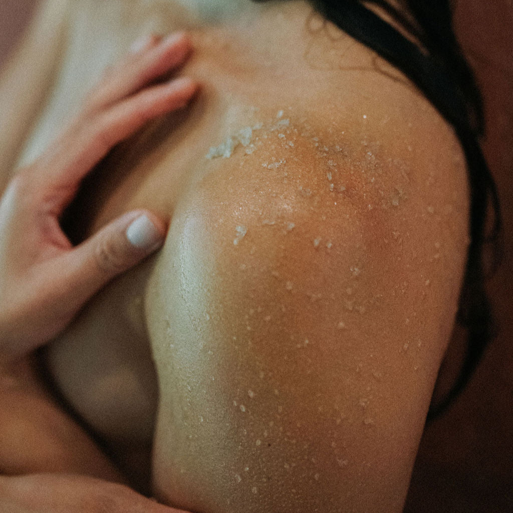 Close-up of a person's shoulder with water droplets and a hand applying a body scrub to it.