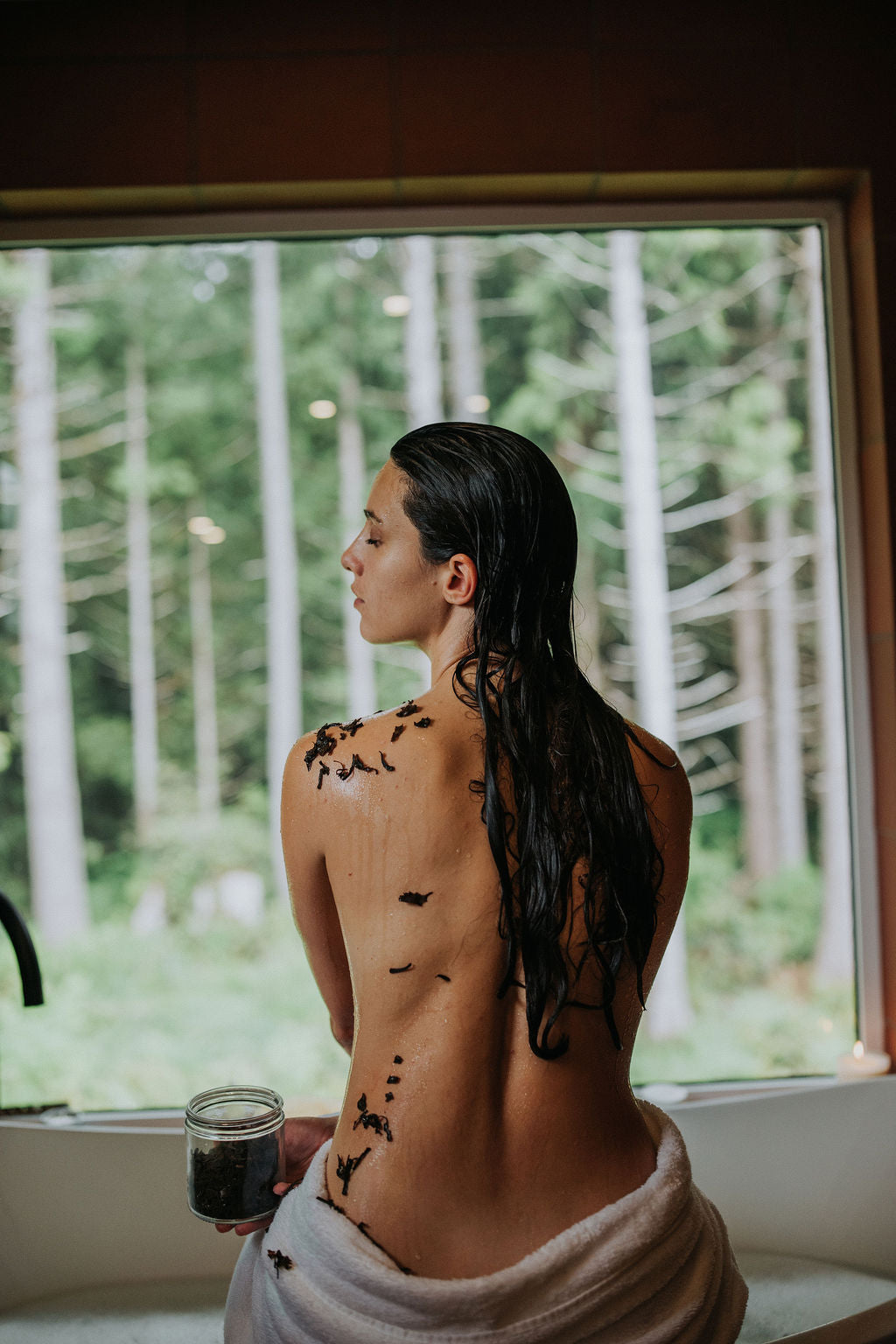 Woman with pieces of seaweed on her back standing in front of a window with a forest view.
