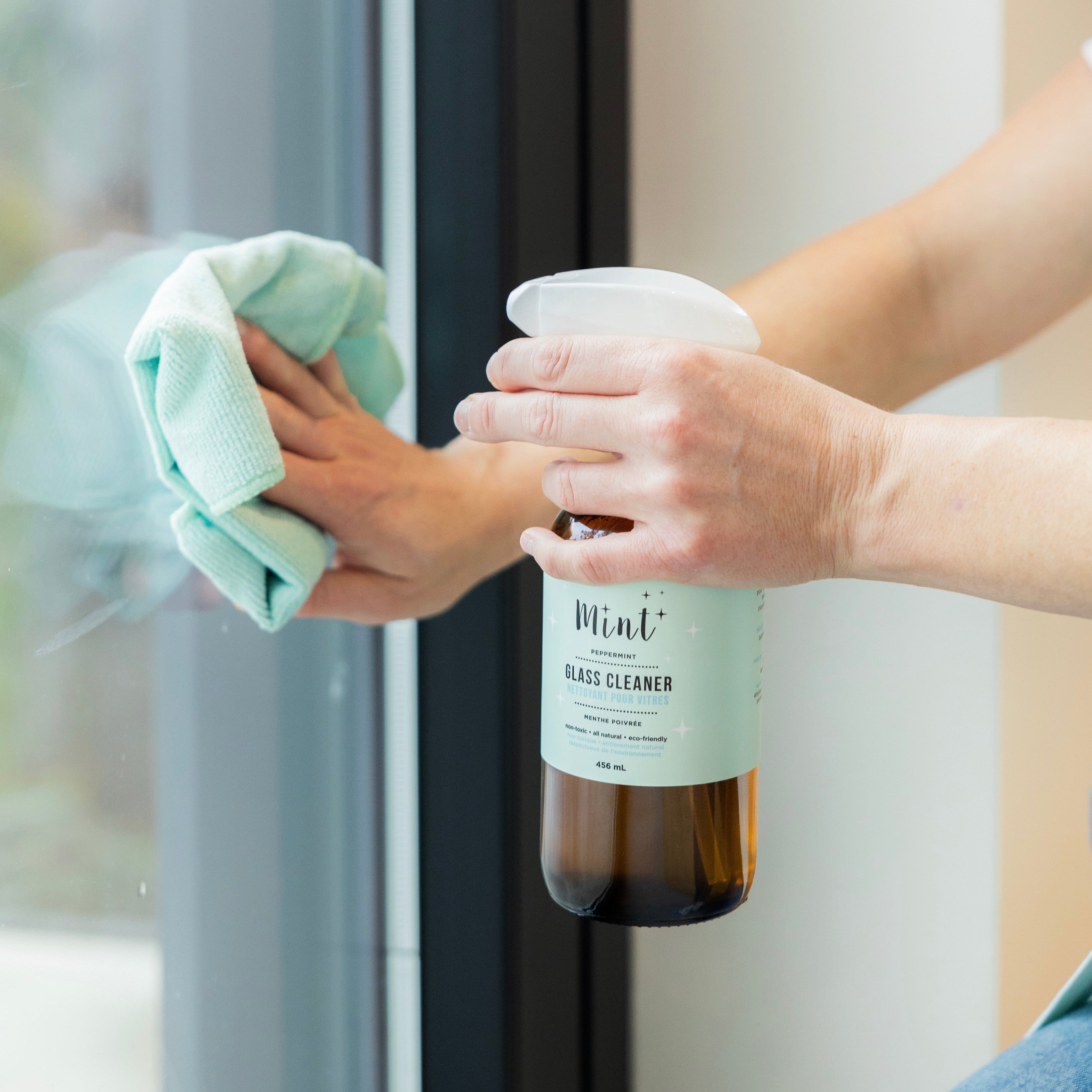 Mint glass cleaner in an amber bottle and white spray cap being used to clean a window. Someone is spraying it onto the surface and wiping it away with a blue microfibre towel
