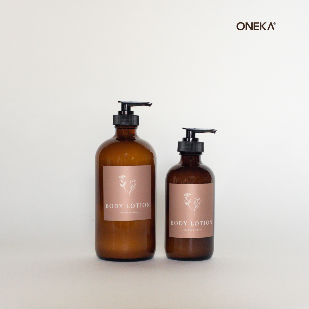 Two amber glass bottles filled with ONEKA lavender and Angelica body lotion sit in a white set. The larger [left] bottle is 500ml, and the smaller [right] bottle is 250ml