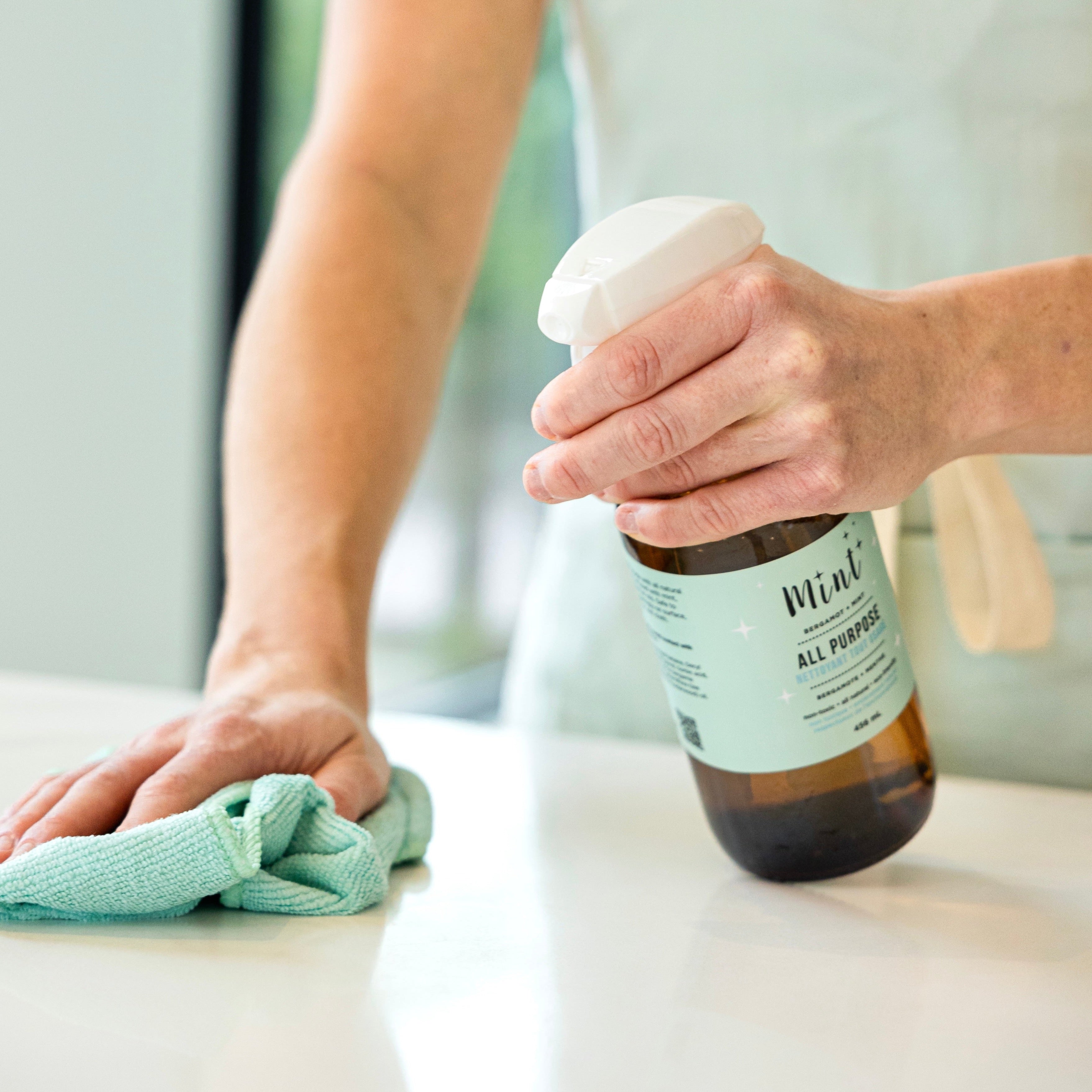 The Mint Cleaning Co. All Purpose Cleaner is being used to clean a white counter top. An individual is spraying the surface and wiping it up with a blue microfibre towel. The amber glass bottle has the classic Mint colour label. 