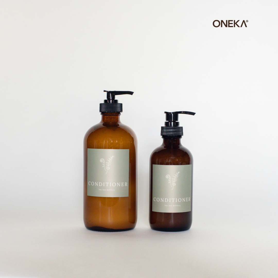 ONEKA cedar and sage conditioner in the Den Refillery’s signature glass amber bottles with green conditioner labels that have white lettering and an illustrated fern. There are two sizes: 500ml [left] and 250ml [right].