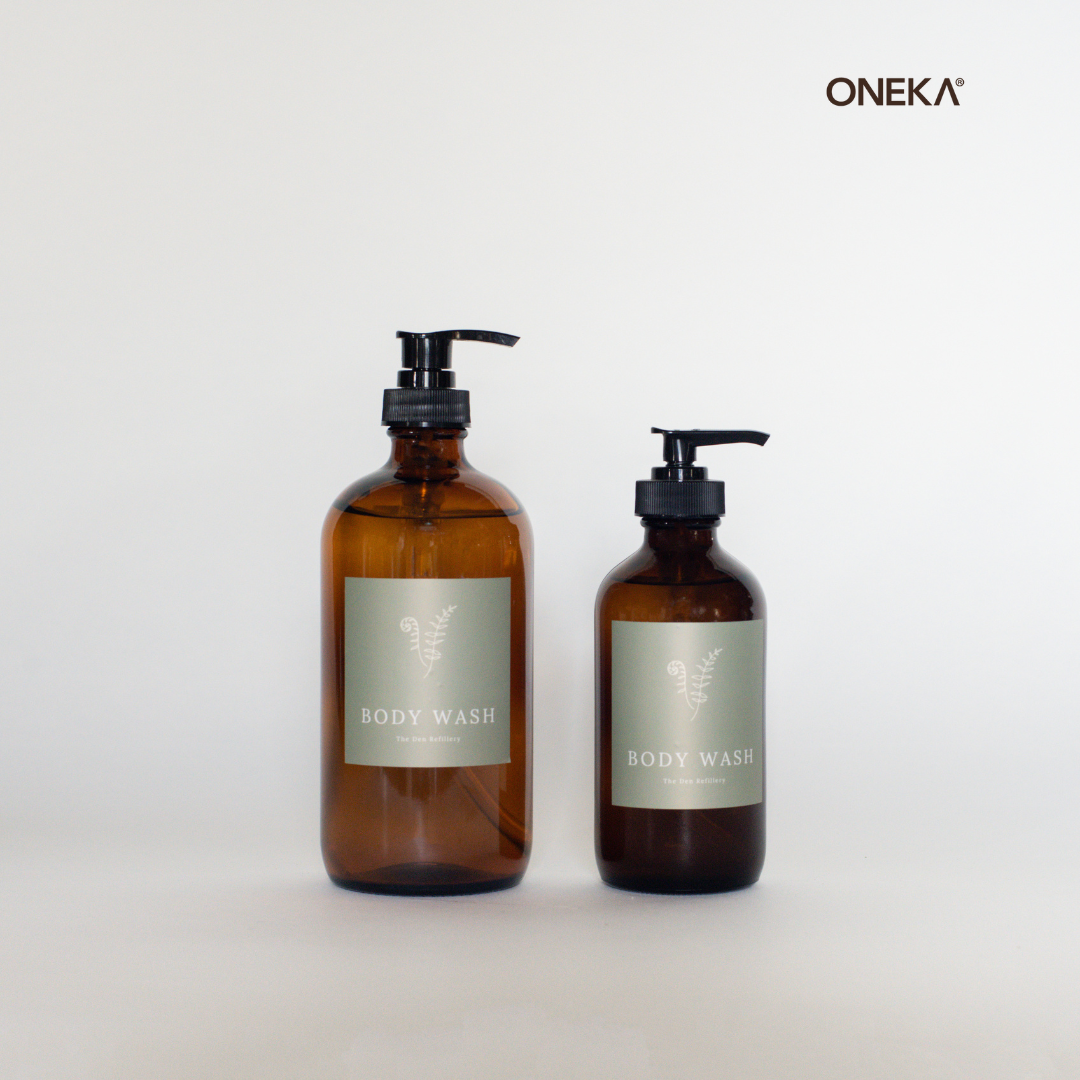 ONEKA Body Wash is available in two signature Den Refillery Amber glass bottles. They sit in a white room with a green Body Wash label