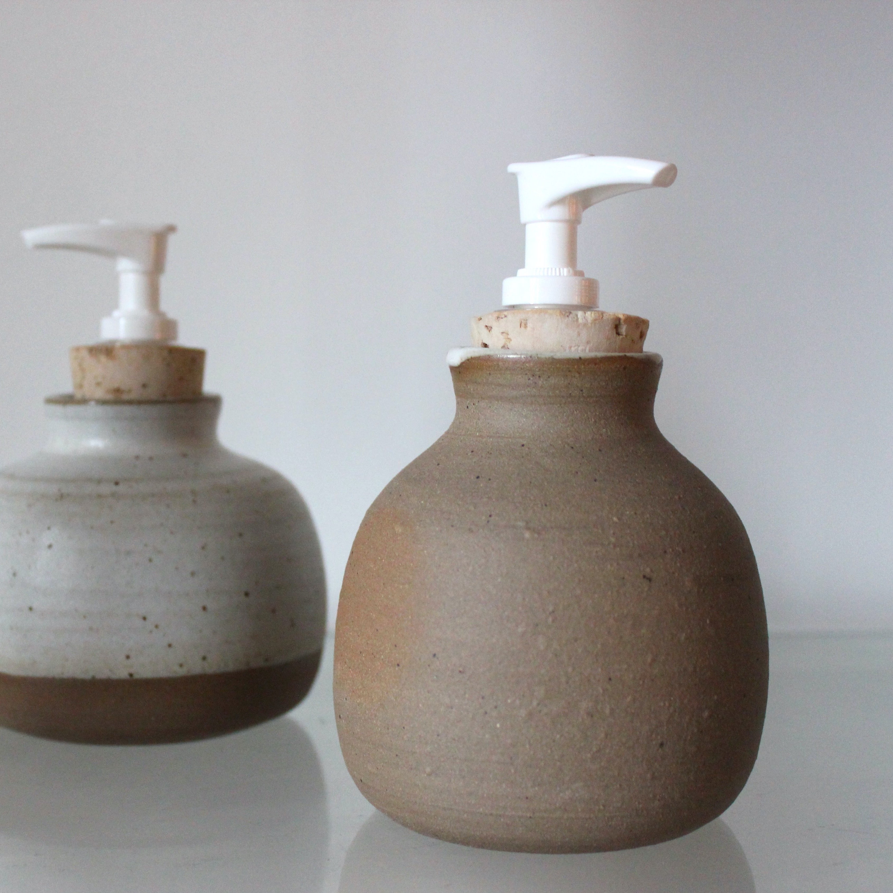 Two of Kay Ceramics ceramic soap dispensers sit on glass. The closer one is sand coloured with a splash of white glaze at the top with a white plastic pump with cork, and the one in behind uses red clay and a white glaze on top with the same plastic and cork pump