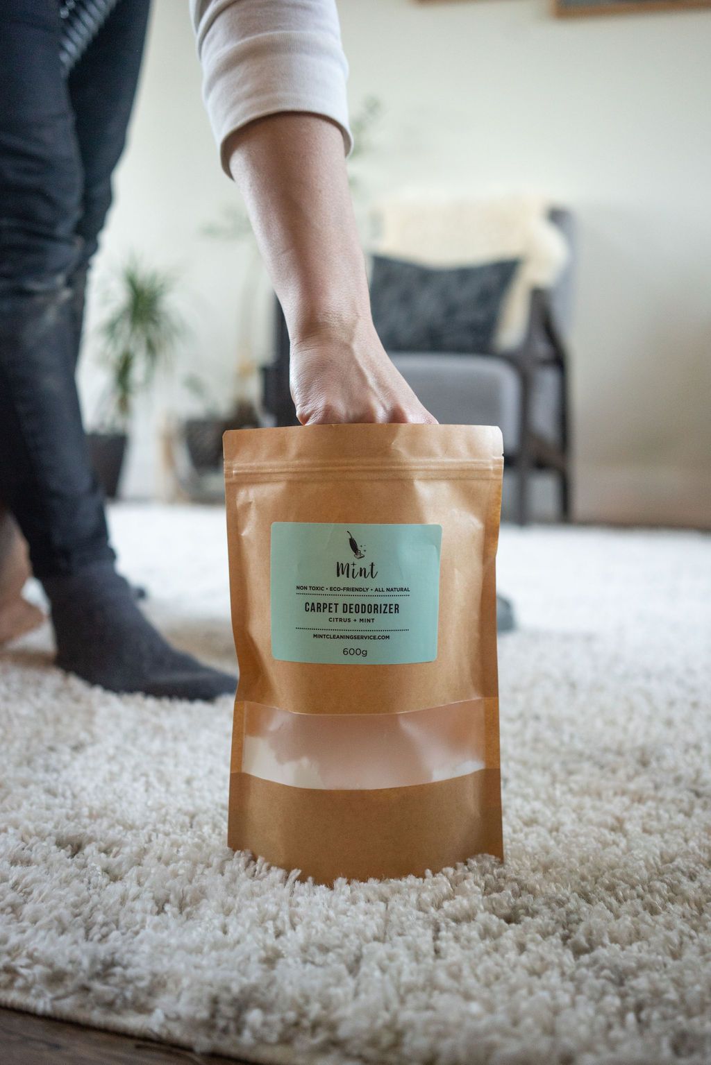 A person is reaching into a paper bag of Mint Cleaning CO’s Carpet Cleaner. They appear to be cleaning their soft white carpet in their comfy living room.