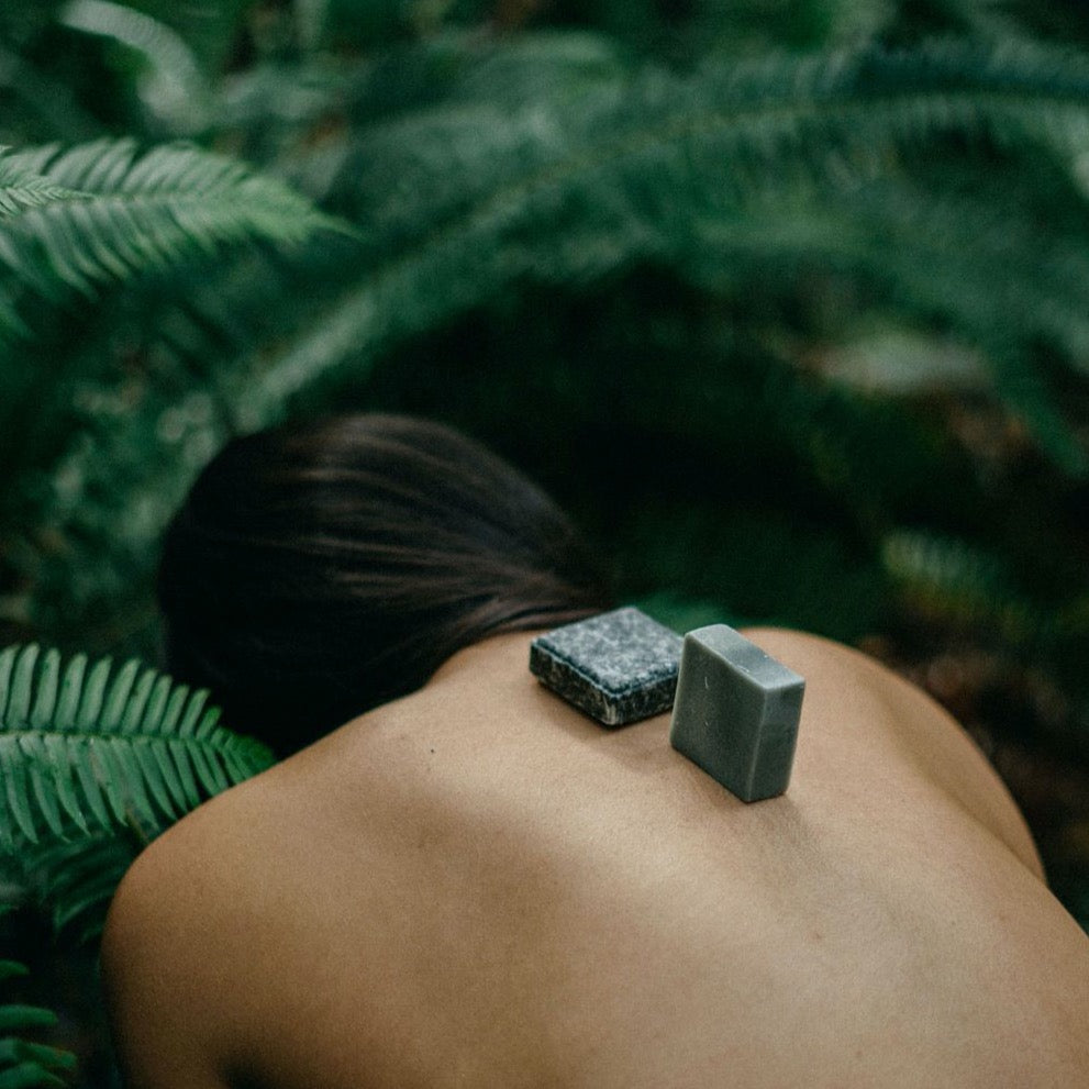 A shampoo and conditioner bar sit on the back of a woman’s bare back as she leans over in the shower
