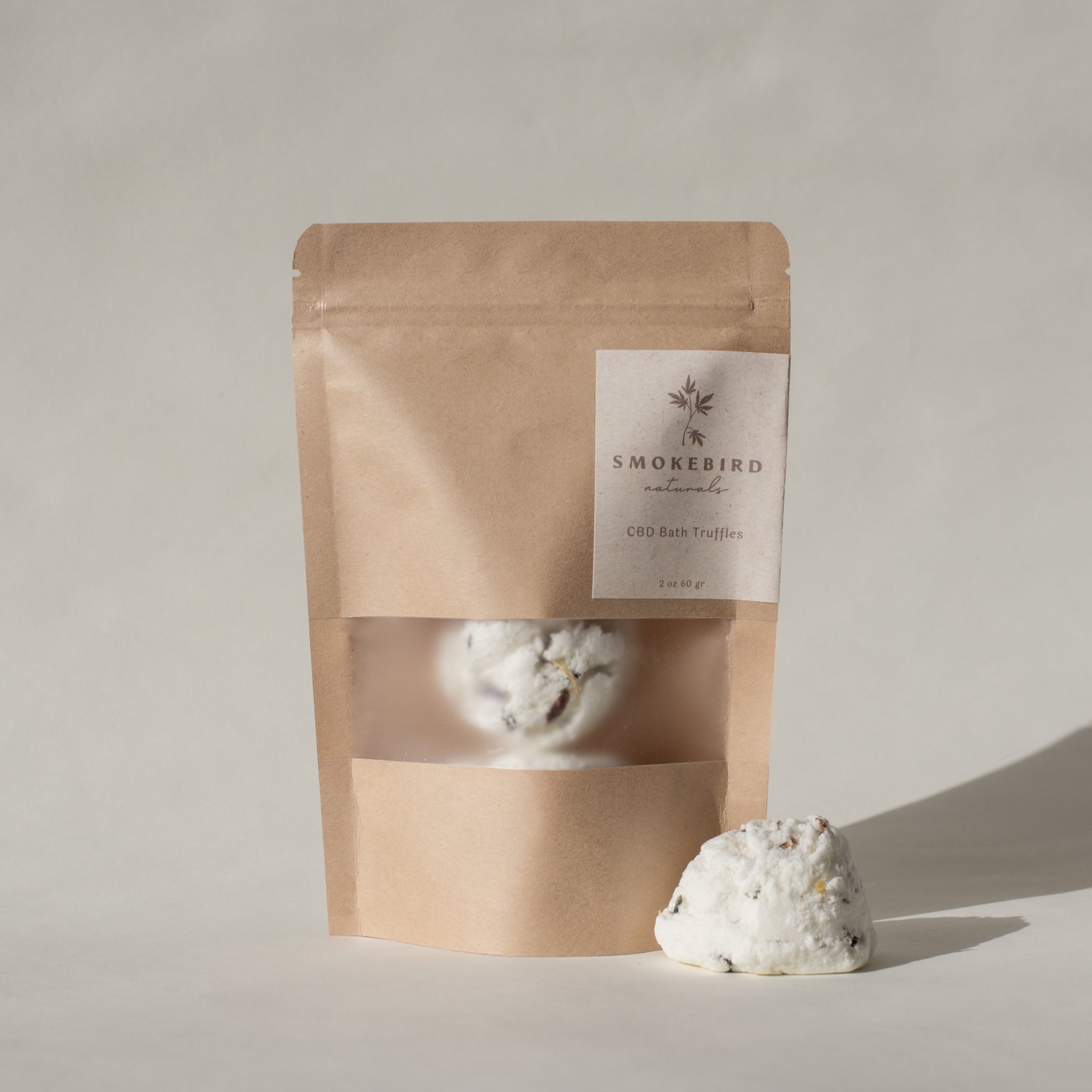 A paper bag of Smokebird Botanicals Bath Truffles sits in a white room with one of the bath truffles out on the table in front of the package